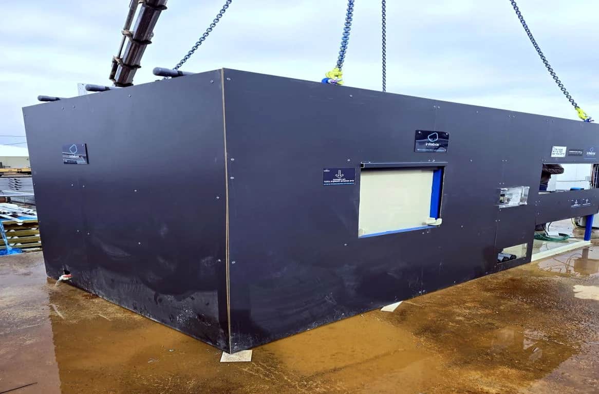 A black INFRABOX unit on crane chains being lowered onto its prepared pad